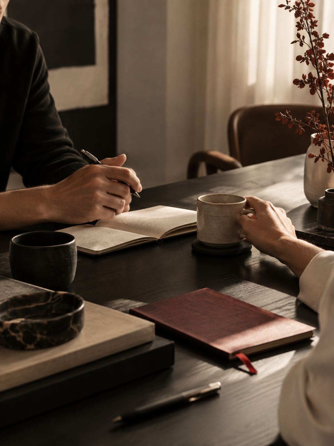 A person writing in a notebook at a dark wooden table, a ceramic mug and a red notebook